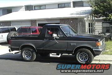 Dark gray 1987 Ford Bronco with black trim and a spare tire on the back. Another SUV is parked behind it