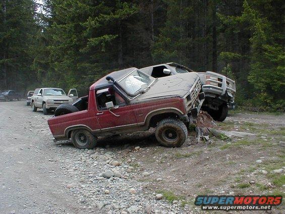 Red SUV stuck on a log with another truck behind it. Other vehicles are parked on the side of the dirt road