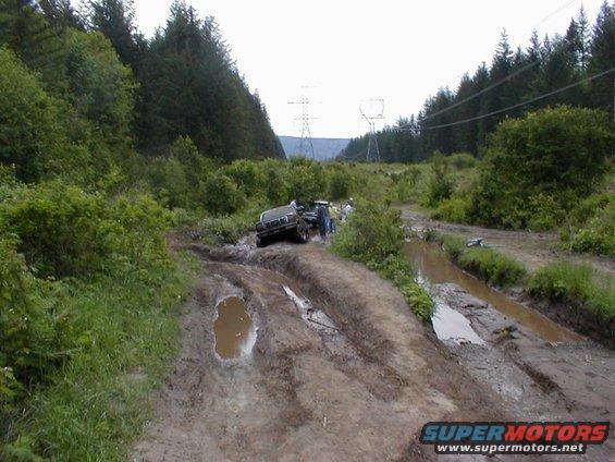 Two off-road vehicles stuck in deep mud on a trail with trees and power lines