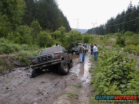 Brown Ford F-Series truck stuck in mud on a trail, with other trucks and people nearby