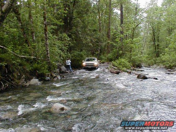 Tan SUV driving through a shallow river, front view. Trees and rocks line the riverbanks