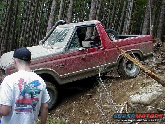 Red Ford Bronco with snorkel off-road in a forest, tilted on a muddy incline
