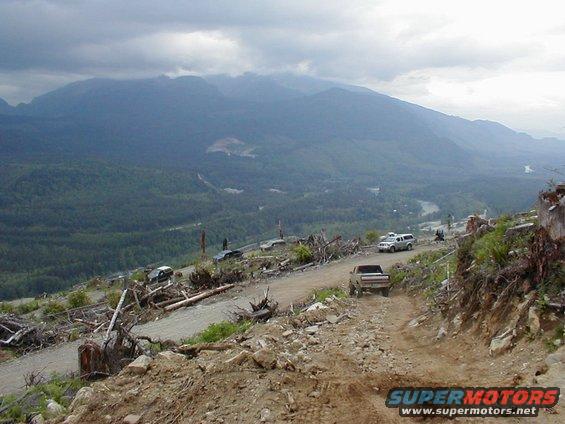 Trucks and SUVs driving on a dirt road up a mountain with trees and debris on the side