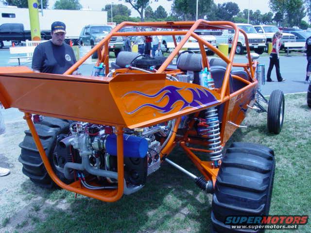 Orange custom buggy with purple flames, large rear tires, exposed engine, and roll cage. A man stands nearby