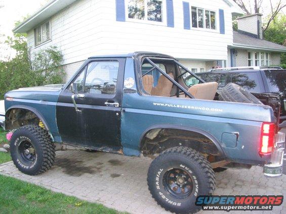 Modified Ford Bronco with roll cage, black doors, and large tires. A dark SUV is parked behind it