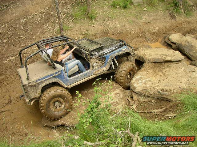 Mud-covered blue Jeep Wrangler navigating large rocks on an off-road trail