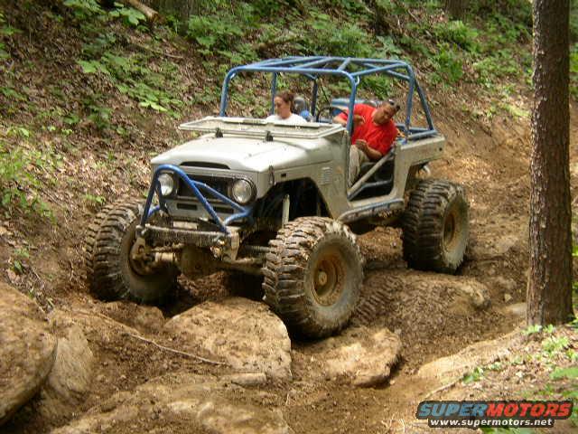 White off-road vehicle with blue roll cage on a rocky, wooded trail