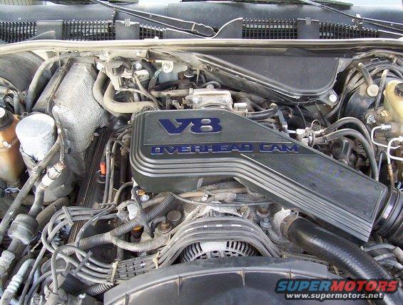 Engine bay with V8 OVERHEAD CAM cover. Watermark reads SUPERMOTORS