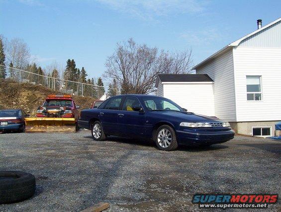 1995 Ford Crown Victoria parked on gravel lot near a white building, with a snow plow truck in the background