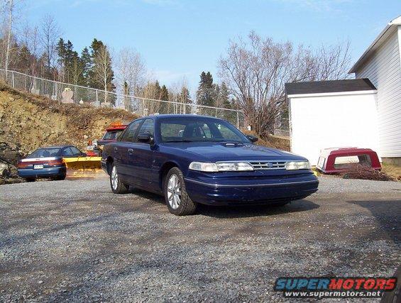 Blue 1995 Ford Crown Victoria parked outside a white building. Another car and a snowplow are visible behind it