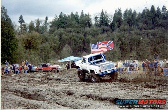 White off-road truck with blue roll cage driving through mud, American and Confederate flags flying