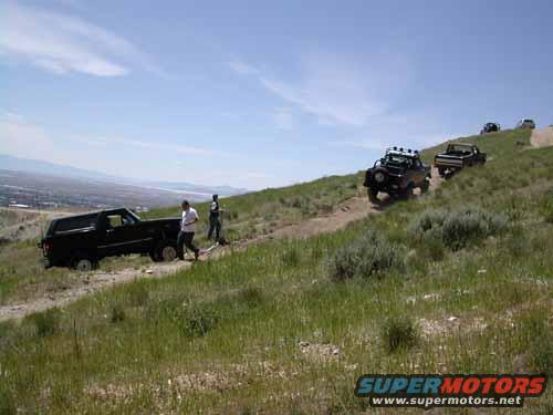 Several off-road vehicles driving up a grassy hill on a dirt track under a blue sky