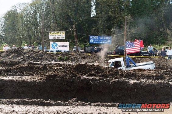 White pickup truck stuck in mud pit, American flag on pole behind it. Other vehicles and spectators visible