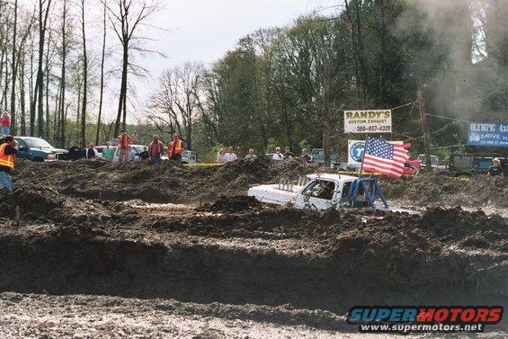 White pickup truck with roll cage stuck in mud, American flag on back. Spectators and trees in background