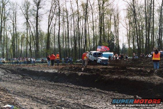 Mud bogging event with trucks stuck in mud, spectators watching, and trees in background. American and Confederate flags on one truck