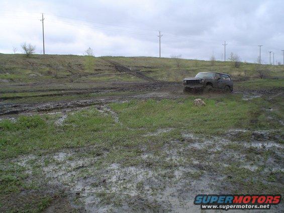 Dark colored 1980 Ford Bronco driving off-road through muddy terrain