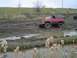 1981 Ford Bronco - Red Chevrolet K5 Blazer navigates a muddy off-road course with other vehicles in the background.