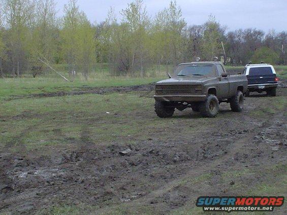 Muddy pickup truck with lifted suspension and off-road tires, followed by a dark SUV on a wet, grassy field