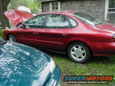 Red sedan with hood open, parked on grass next to a teal car. The red car has a dented rear bumper and aftermarket wheels