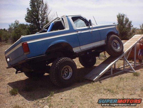 Blue 1987 Ford Bronco with white stripes on a ramp, showing its side and rear
