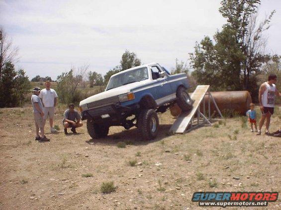 Blue and white pickup truck with lifted suspension driving up a wooden ramp. Three men stand nearby, and a child watches