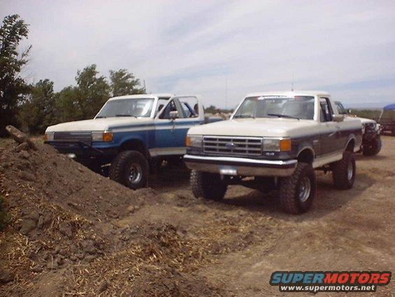 Two lifted Ford trucks, one white and blue, the other white and dark blue, parked on dirt