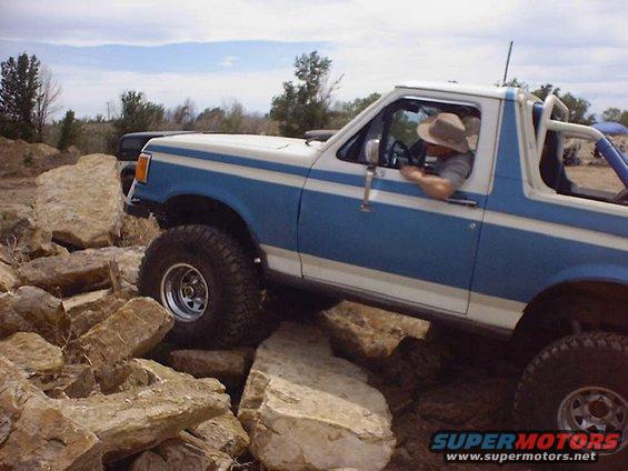 Blue and white 1988 Ford Bronco climbing over rocks, driver wearing a hat