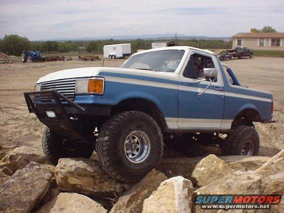 Blue and white Ford Bronco with off-road tires and bumper climbing over large rocks