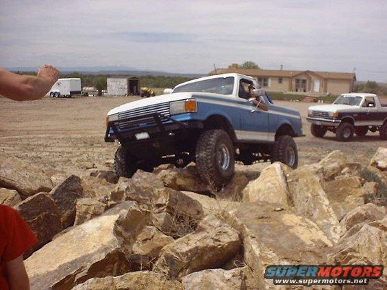 Blue and white pickup truck driving over rocks, another pickup truck behind it