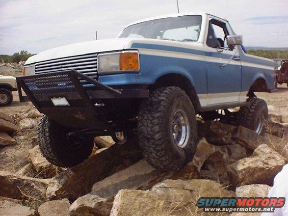 Blue and white Ford Bronco with aftermarket bumper and large tires climbing over rocks