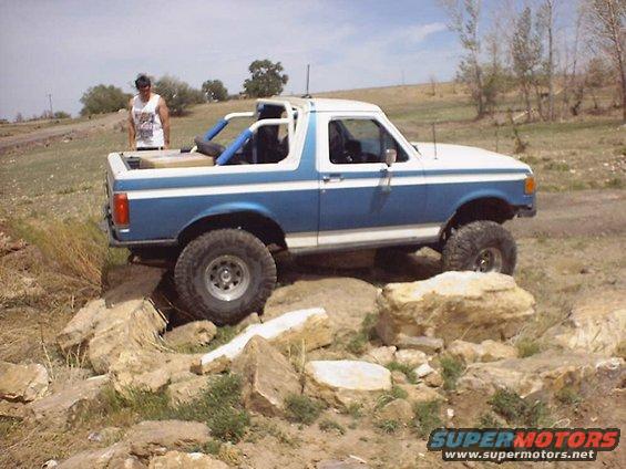 Blue and white Ford Bronco with roll cage, lifted suspension, and off-road tires navigating a rocky trail