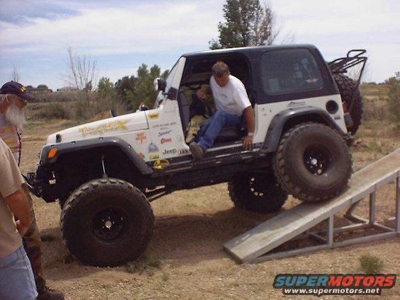 White Jeep Wrangler with black top and off-road tires on a metal ramp, driver entering from the side
