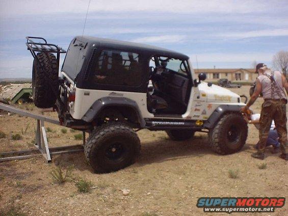 White Jeep Wrangler with black top, spare tire on rear rack, and a man working on the front wheel