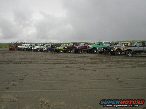 Several trucks and SUVs parked in a line on a sandy beach under a cloudy sky