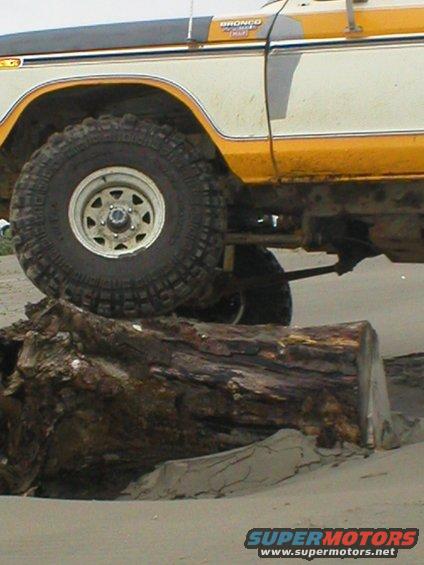 White and yellow 1978 Ford Bronco with off-road tires on a beach, front wheel over a log