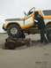 1978 Ford Bronco - A man enters a yellow and white 1978 Ford Bronco parked on a sandy beach, with the front tire resting on a large log.