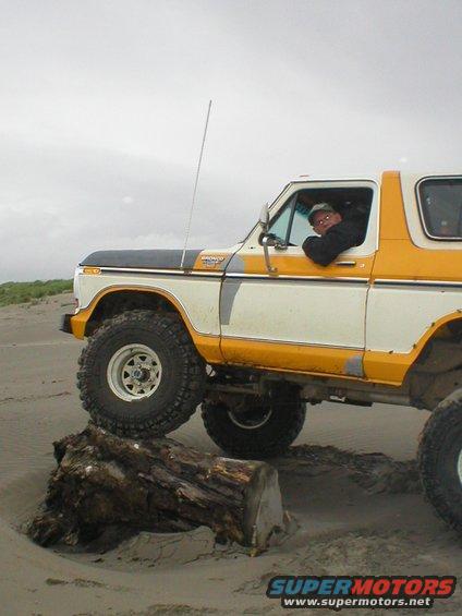 Ford Bronco with yellow and white paint, lifted suspension, driving over a log on a sandy beach. Driver visible