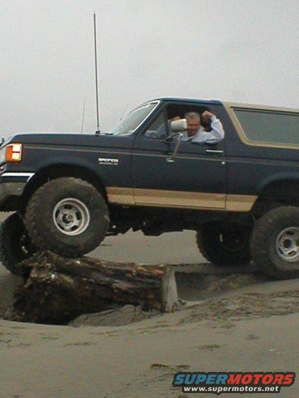 Dark blue SUV with tan top, lifted suspension, driving over a log on a beach. Appears to be a 1990s Ford Bronco