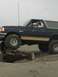 1978 Ford Bronco - A man drives a dark blue SUV with a tan top over a log on a sandy beach, demonstrating its off-road capability.