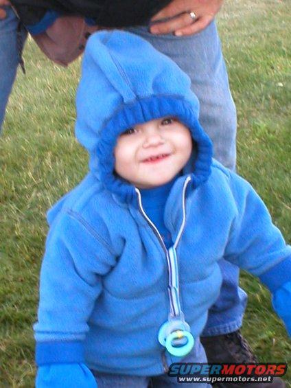 Baby wearing a blue hooded jacket and mittens, smiling outdoors