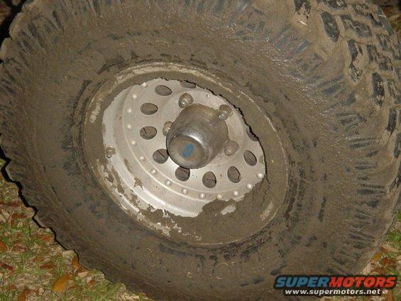 Mud-covered off-road tire with a silver multi-hole wheel and chrome hubcap. The tire tread is deeply grooved