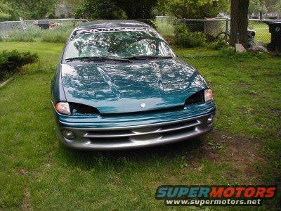 Green sedan parked in a grassy yard, missing both headlights. A banner is visible across the top of the windshield