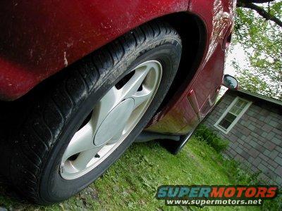 Close-up of a red car's rear wheel and fender. A brick building is visible in the background