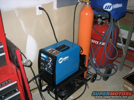 Blue Miller welding machine next to orange gas cylinder and red air compressor