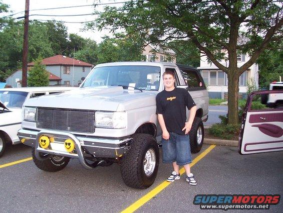 Silver lifted Ford Bronco with custom grille and smiley face fog lights parked next to a white car and a car with open doors