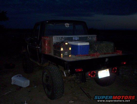 Flatbed Ford truck at night, loaded with a toolbox, cooler, and other gear. License plate is visible