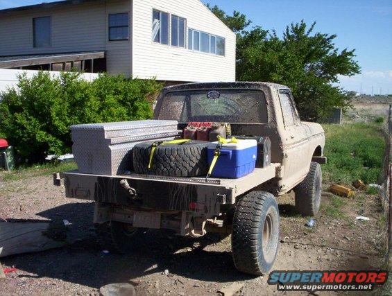 Rear view of a tan flatbed truck with a toolbox, spare tire, and cooler