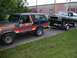 1979 Ford Bronco - A black and orange Ford Bronco tows a green Ford Bronco on a trailer down a paved driveway.