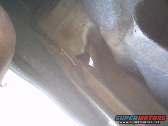 Underside of a car, showing frame and some suspension components. A person's arm is visible on the left