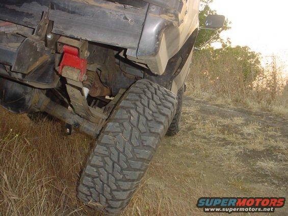 Rear leaf spring suspension with red shackle flip bracket and large off-road tire on a dirt track